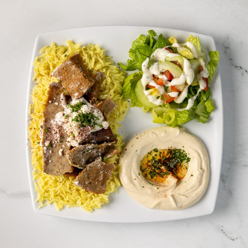 Mediterranean platter with shawarma, yellow rice, hummus, creamy-dressed salad, and pita on a white plate.