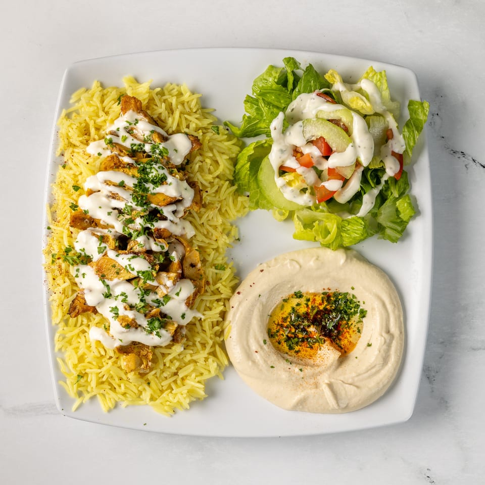 Mediterranean platter with yellow rice, grilled chicken drizzled with sauce, hummus, and dressed lettuce salad.