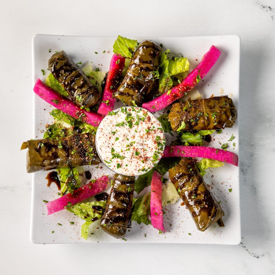 Stuffed grape leaves with bright pink pickled radishes, lettuce, herbs, and creamy dip on a white plate.