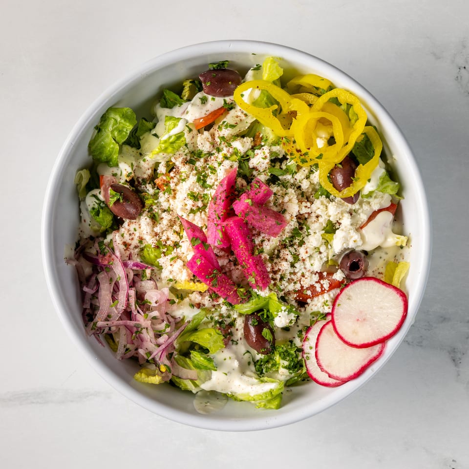 Overhead view of a colorful Mediterranean salad bowl with lettuce, feta, olives, peppers, pickled vegetables, radishes, and creamy dressing.
