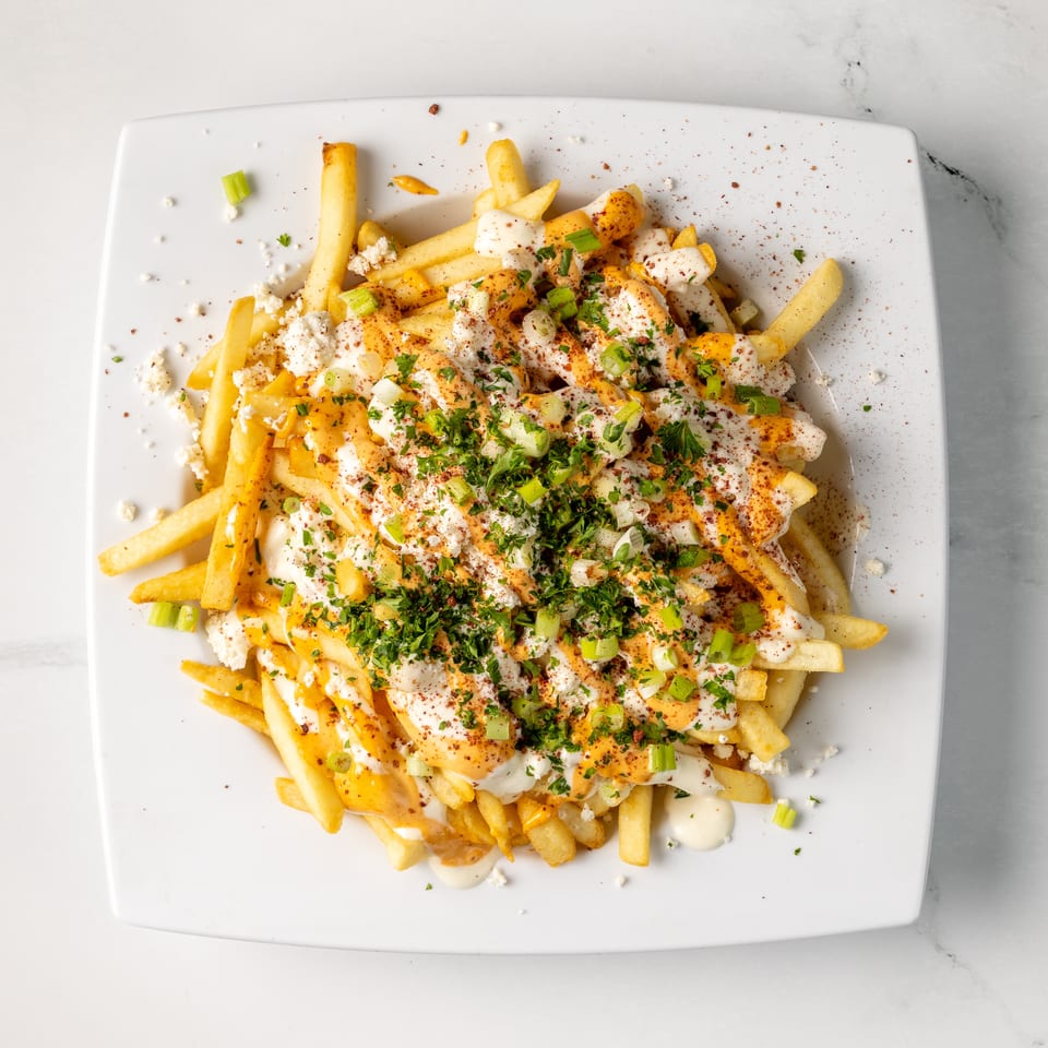 Loaded French fries topped with creamy sauces, crumbled cheese, chopped herbs, green onions, and spices on a white plate.