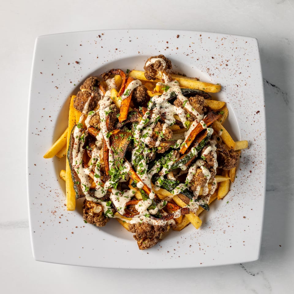 Loaded fries topped with roasted vegetables, crispy cauliflower, creamy sauce, herbs, and spices on a white plate.
