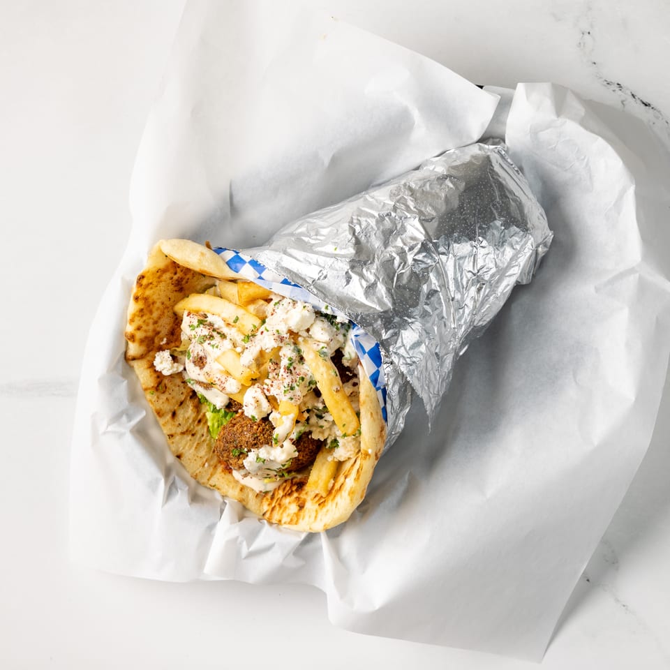 Open pita sandwich filled with falafel, fries, lettuce, feta, and creamy sauce, partially wrapped in foil on white paper.