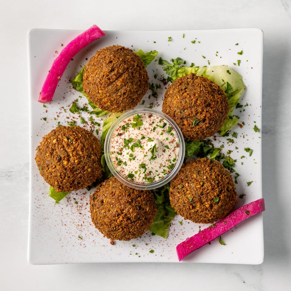 Falafel patties arranged on lettuce with creamy dipping sauce, chopped herbs, spices, and bright pink pickled vegetables.