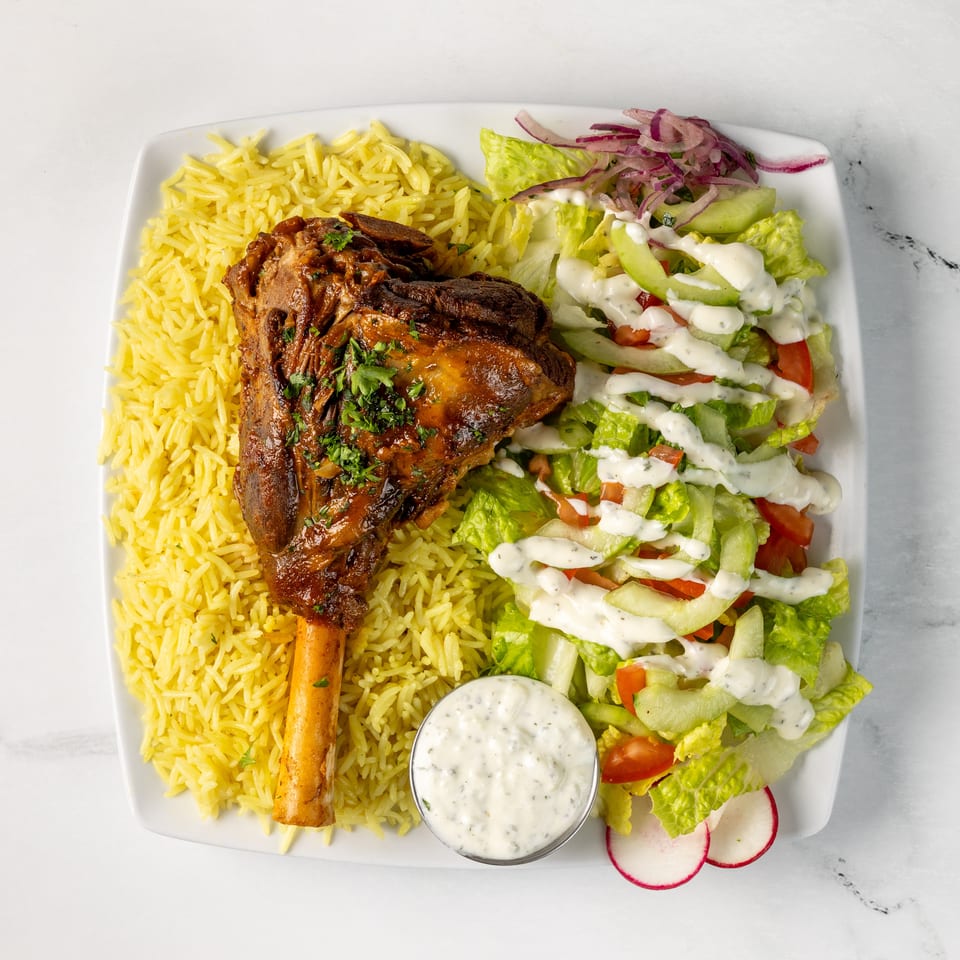 Plate of roasted lamb shank with yellow rice, chopped salad, creamy dressing, and dipping sauce.