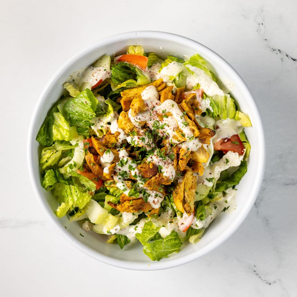 Top-down view of a leafy salad bowl topped with seasoned chicken, tomatoes, cucumbers, creamy dressing, herbs, and spices.